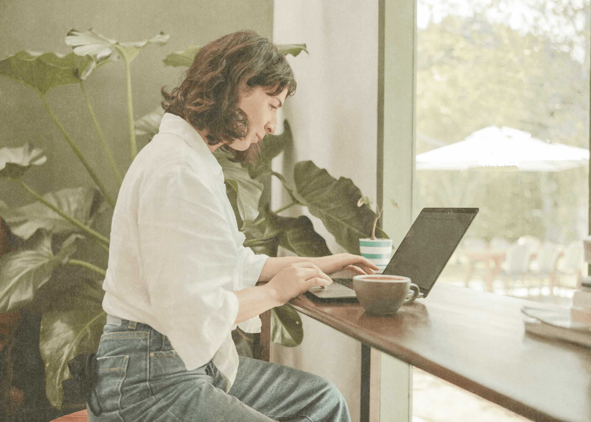 Woman with curly hair working on a laptop at a wooden counter near a large window with a cup of coffee and green plants in the background.